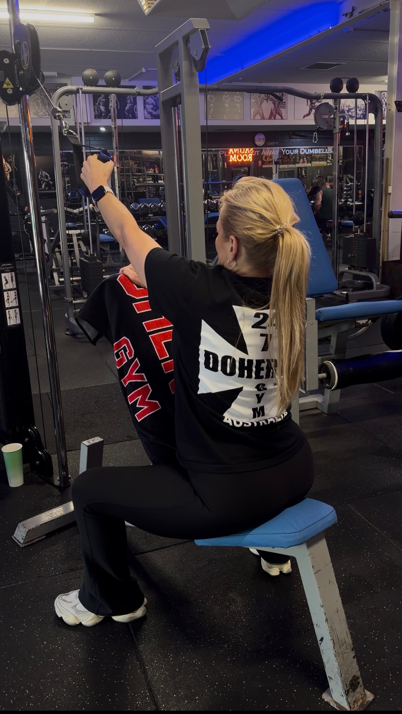 A woman with a blonde ponytail seated, doing a cable pulldown at the gym.