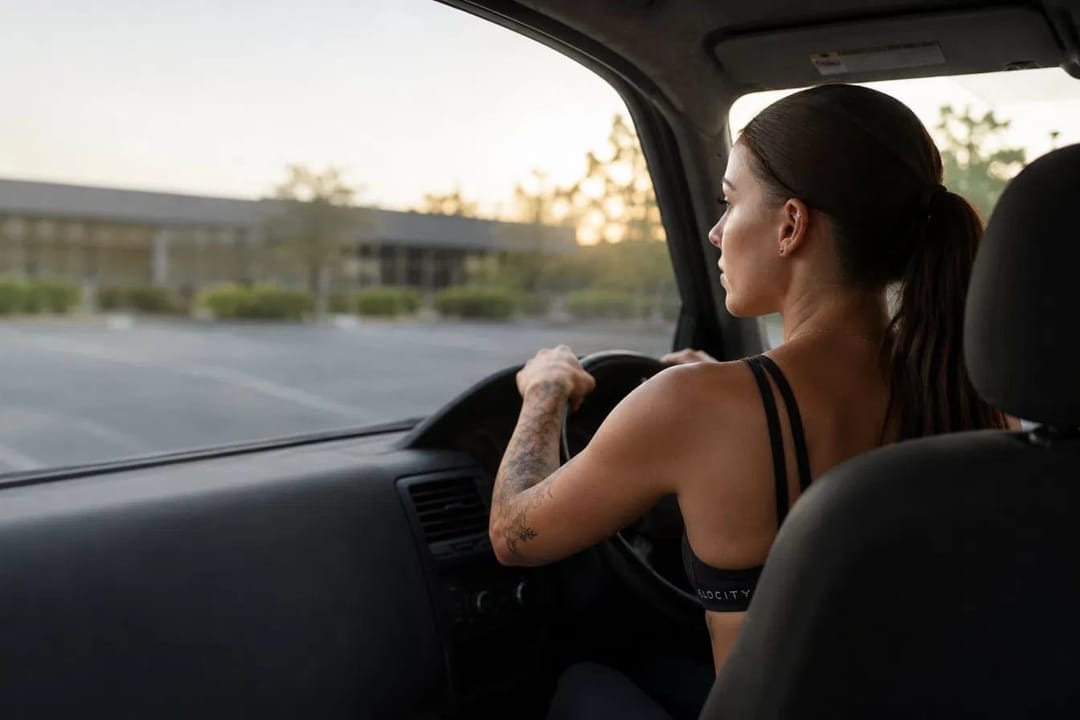 A woman sitting in her car in a gym car park at sunrise, looking out towards the building.